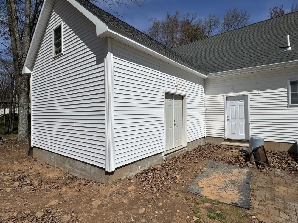 Exterior view of a white-sided house with two doors, a small patio, and surrounding trees under a clear blue sky.