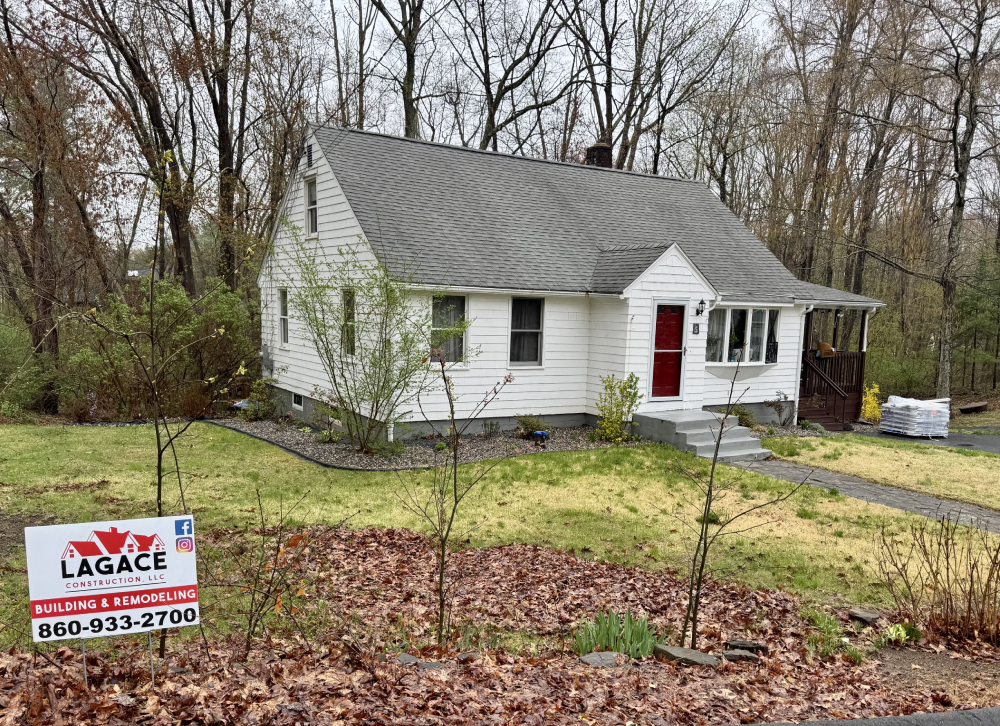 A charming white house with a gray roof, surrounded by trees, featuring a sign for L'Agace Construction, advertising building and remodeling services.