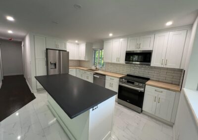 Modern kitchen with white shaker cabinets, light wood countertops, and stainless steel appliances. Features a black countertop island, subway tile backsplash, and recessed lighting. The kitchen has a clean, bright design with white marble-look tile flooring. Located in Andover, Connecticut.