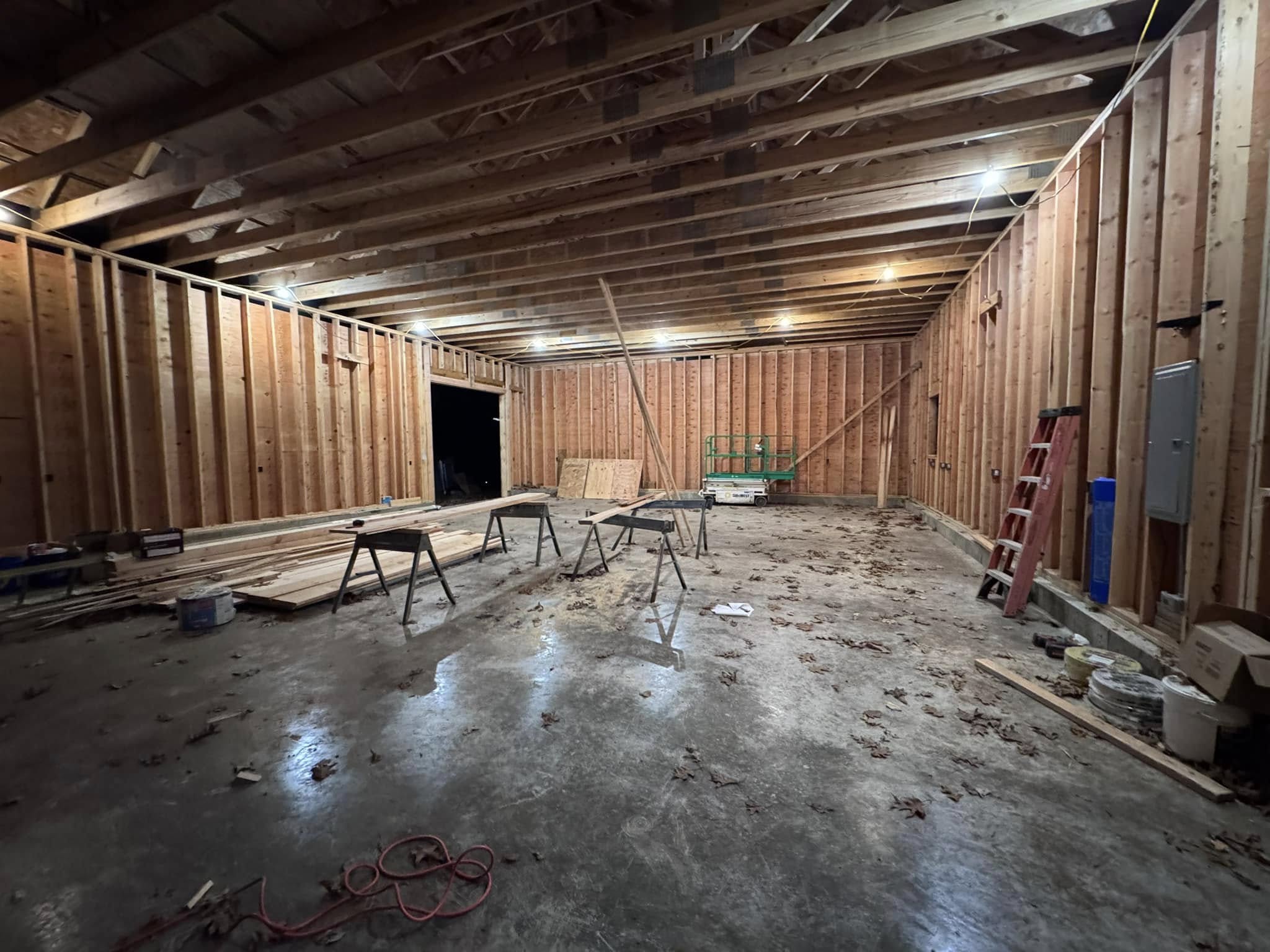 Large unfinished interior space with exposed wooden framing and open ceiling joists, prepared for remodeling. The area features a concrete floor, scattered construction tools, sawhorses, a red ladder, and stacked lumber. Overhead work lights illuminate the structure. Located in Columbia, Connecticut.